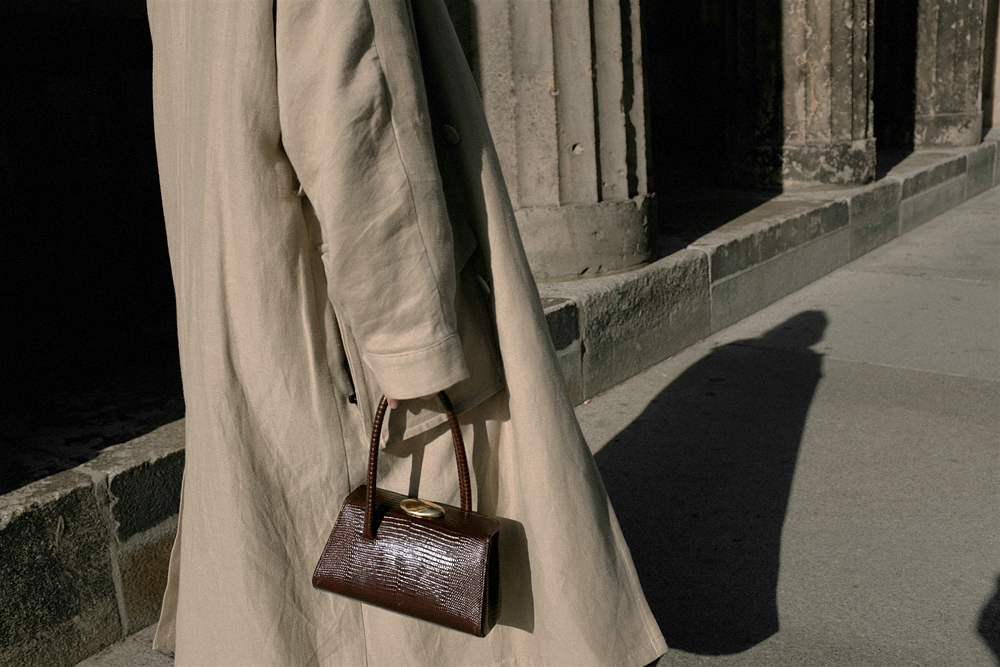 Nisi is wearing: Black wide leg denim jeans – Edited the label, White alpaca-blend sweater – Filippa K, Trenchcoat – & Other Stories, Baby Boss bag – Little Liffner, Heeled flip-flops – Totême, Tire earrings – All Blues