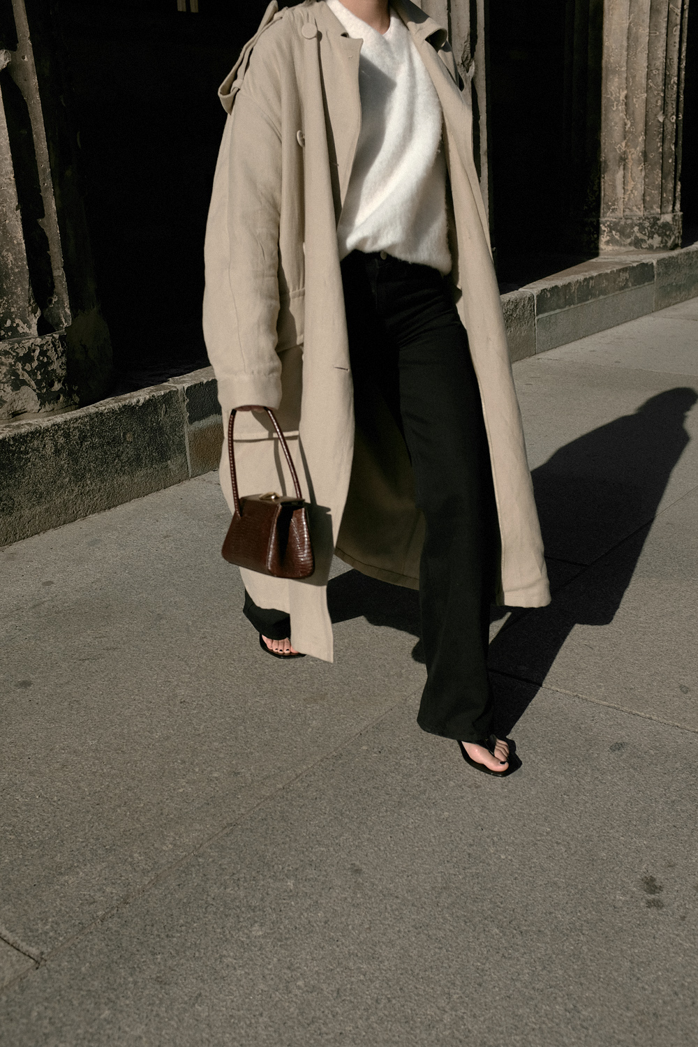 Nisi is wearing: Black wide leg denim jeans – Edited the label, White alpaca-blend sweater – Filippa K, Trenchcoat – & Other Stories, Baby Boss bag – Little Liffner, Heeled flip-flops – Totême, Tire earrings – All Blues