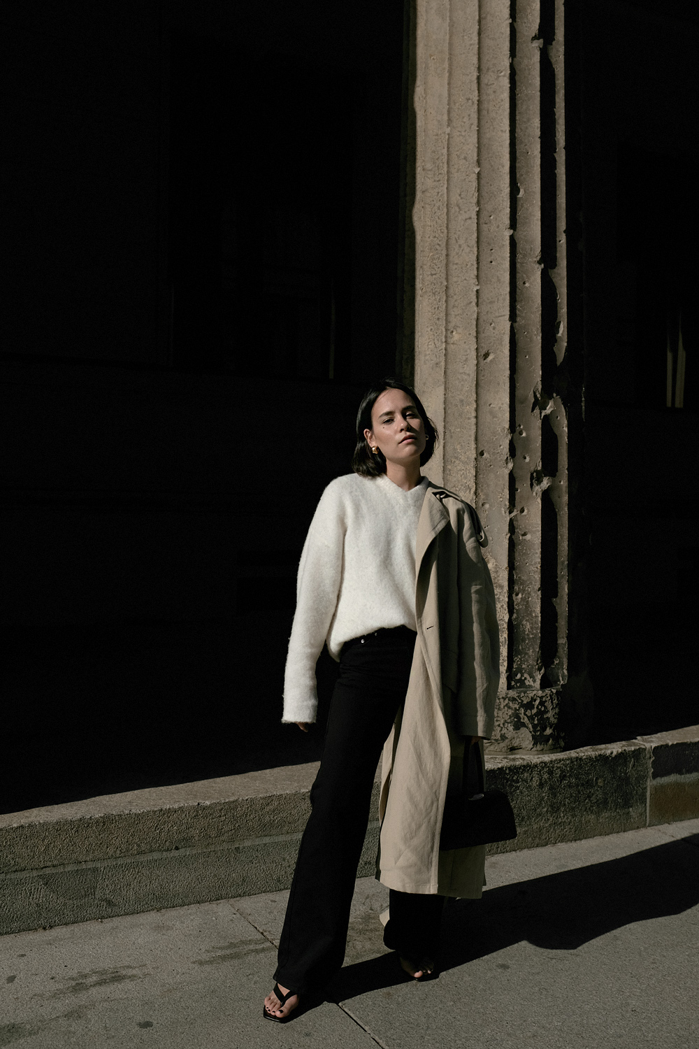 Nisi is wearing: Black wide leg denim jeans – Edited the label, White alpaca-blend sweater – Filippa K, Trenchcoat – & Other Stories, Baby Boss bag – Little Liffner, Heeled flip-flops – Totême, Tire earrings – All Blues